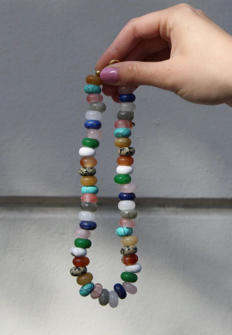 Hand holding a natural stone necklace with colorful, eclectic beads and 18K gold-plated details.